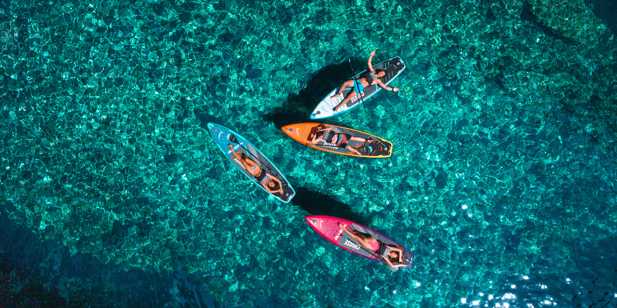 Quattro persone pagaiando su stand-up paddleboard colorati in acque turchesi e cristalline, viste dall’alto