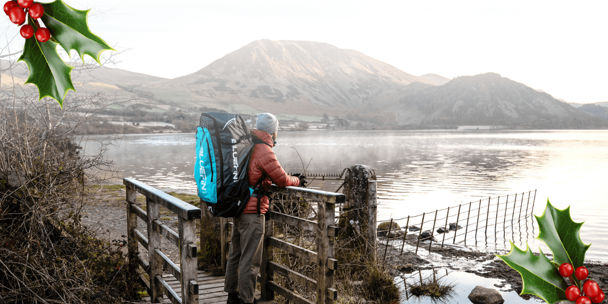 Person wearing a Bluefin backpack stands on a lakeside footbridge beneath snowy mountains, framed by holly leaves and berries