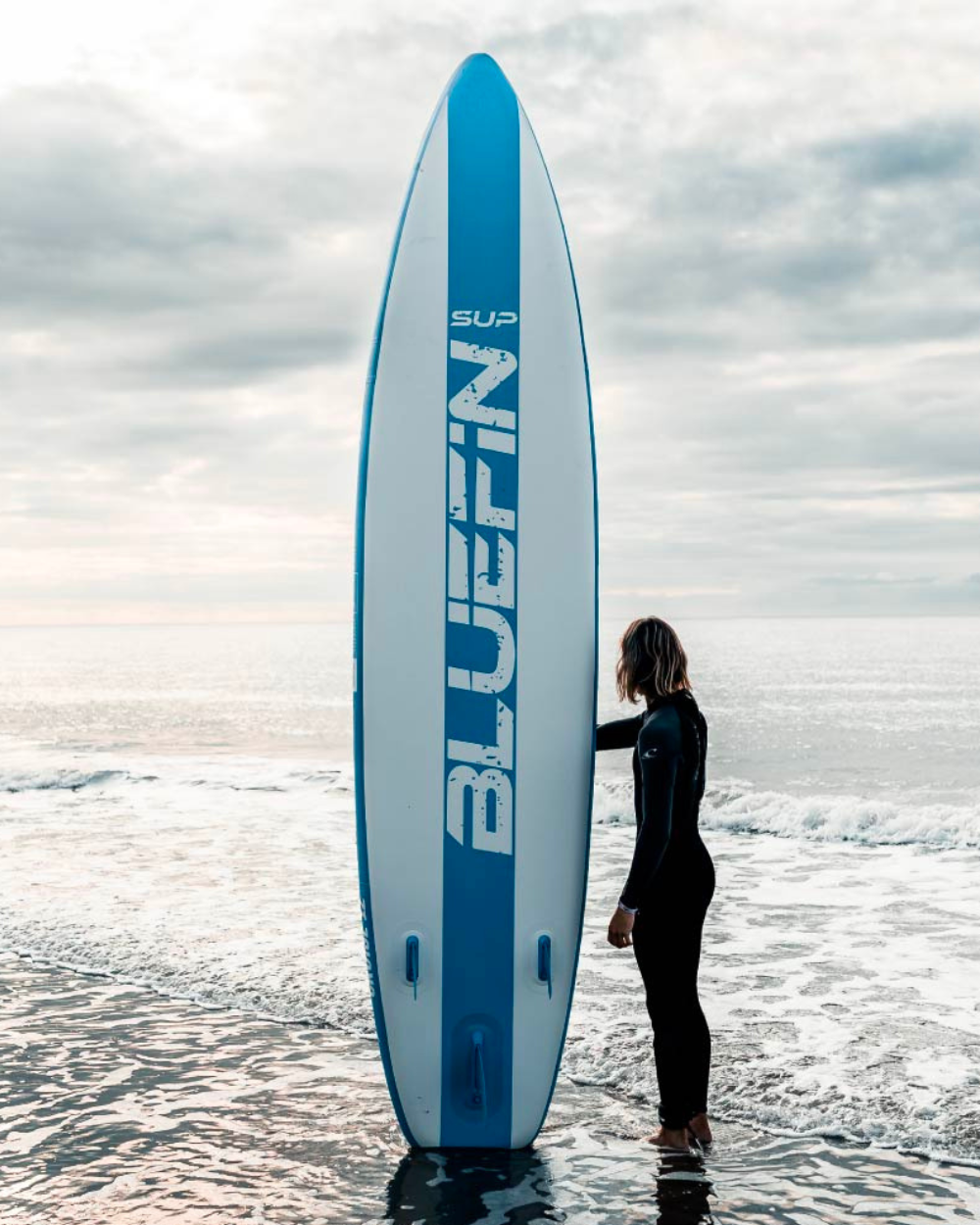 Person in wetsuit standing beside a blue and white Bluefin SUP paddleboard on the shore