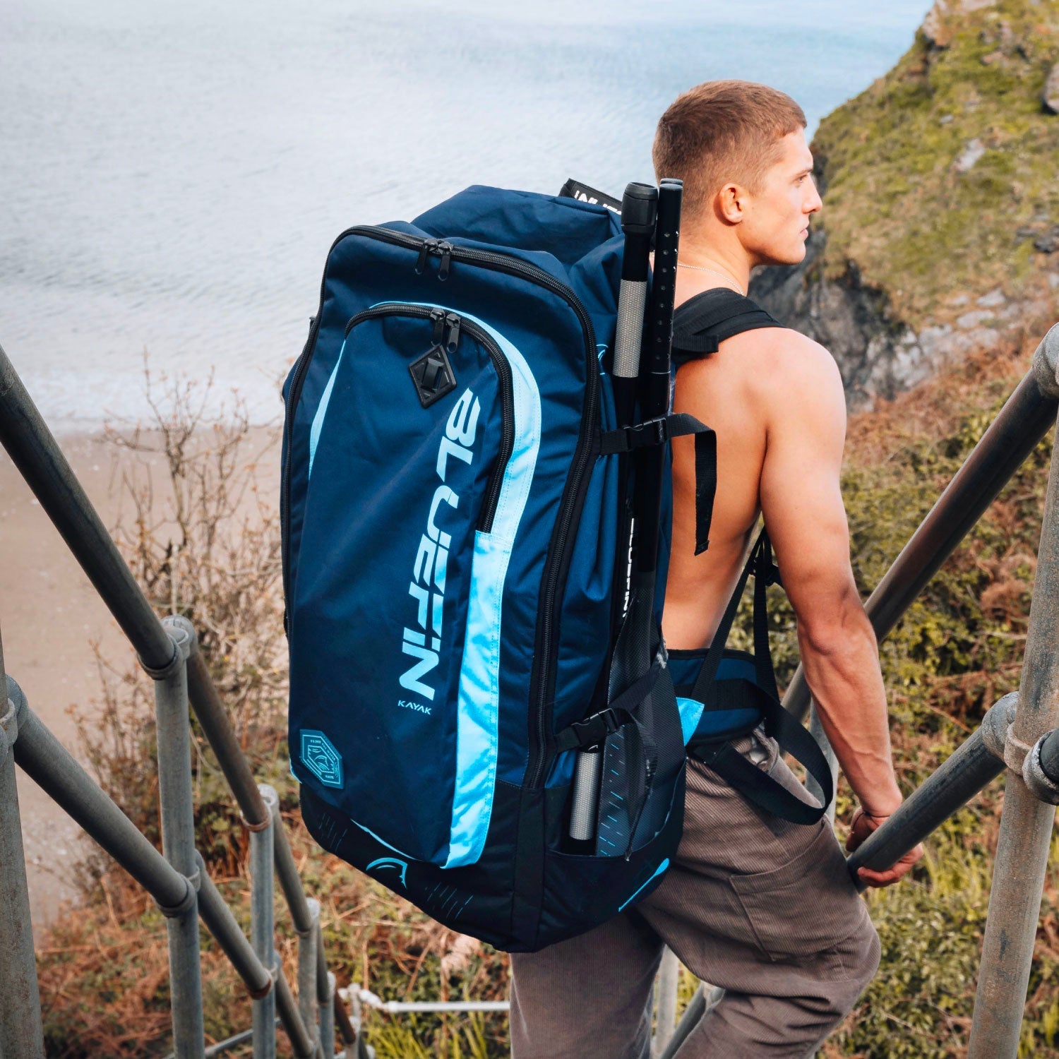 Man carrying a large blue backpack with paddles, walking up metal stairs near a beach.
