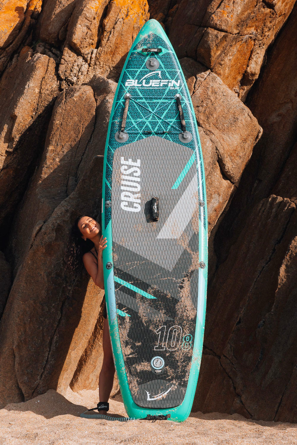 Inflatable paddleboard with a textured deck, center fin, and a woman smiling beside it on the beach.