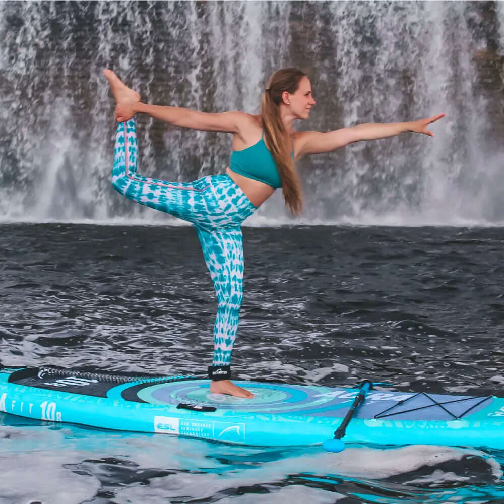 Femme en tenue de sport turquoise pratiquant le yoga sur un paddleboard devant une cascade