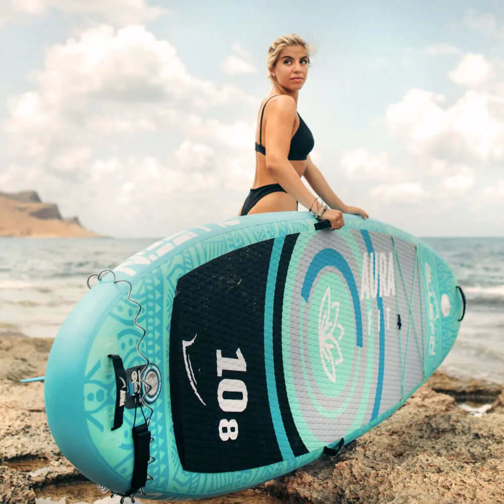 Femme en bikini noir tenant un paddleboard gonflable turquoise au bord de la mer avec des vagues océaniques.