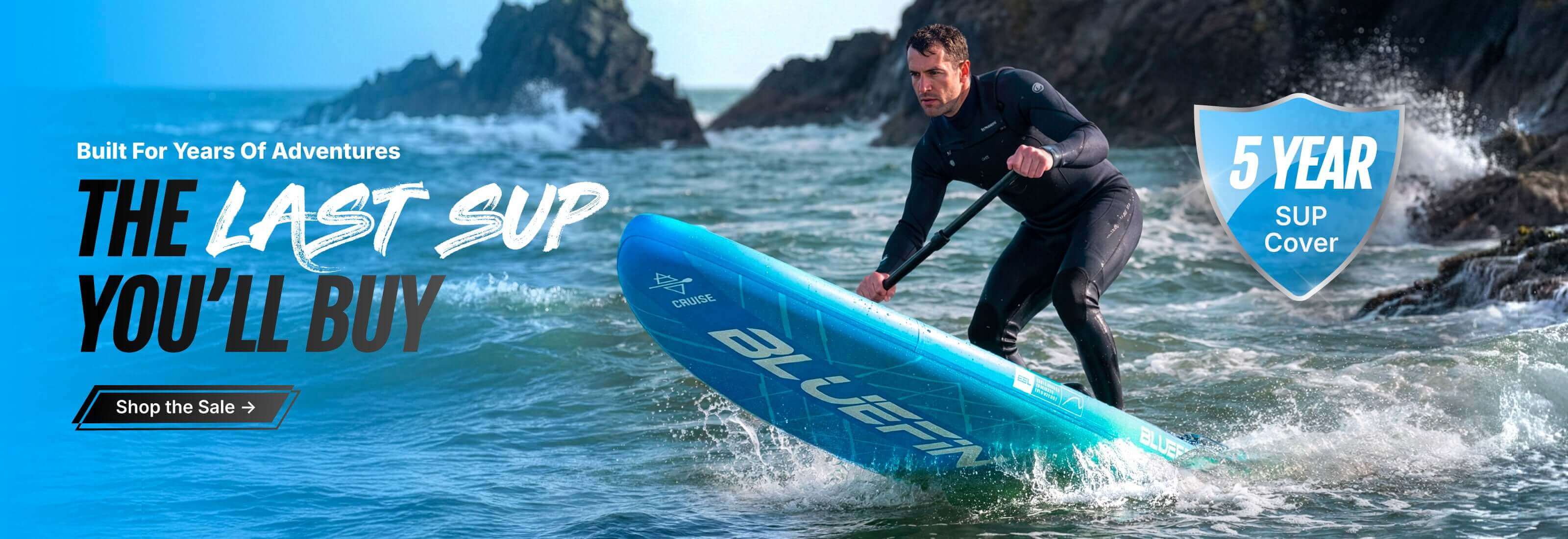 Man in wetsuit paddling a blue inflatable paddleboard on ocean waves near rocky shore