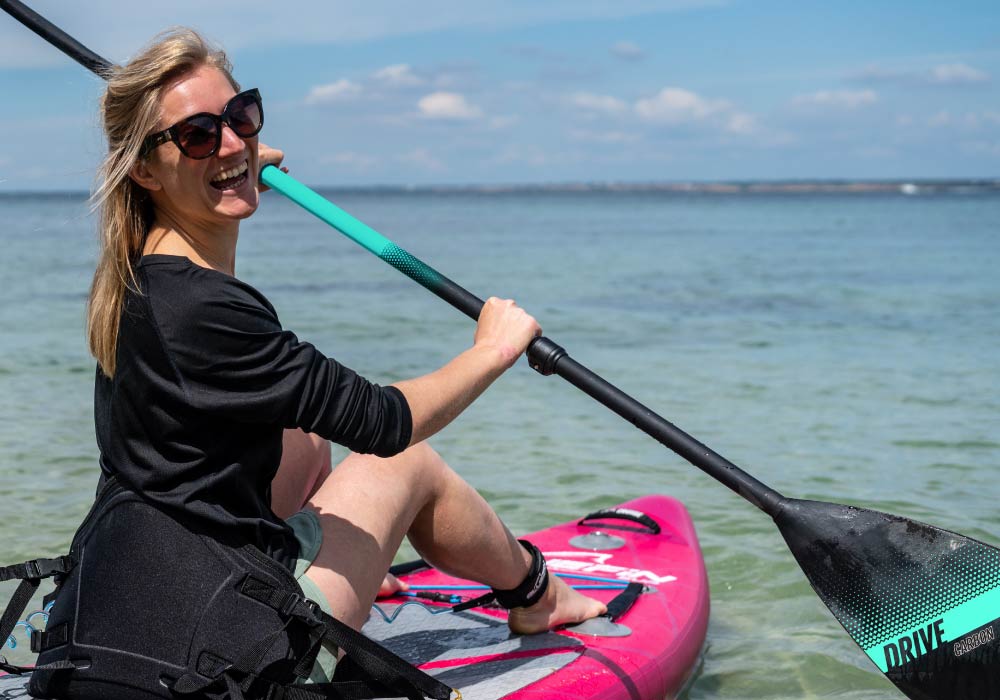 Woman paddling on a pink inflatable paddleboard in clear water, wearing sunglasses and smiling.