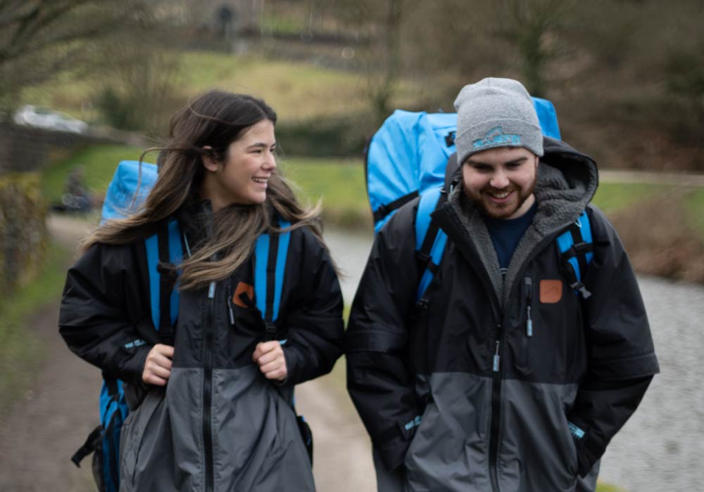 Two hikers walking on a path, wearing black jackets and carrying blue backpacks, smiling at each other.