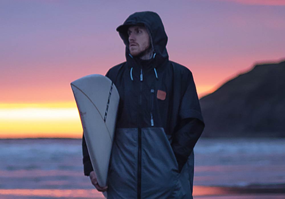 Person in a black jacket holding a surfboard at sunset on the beach