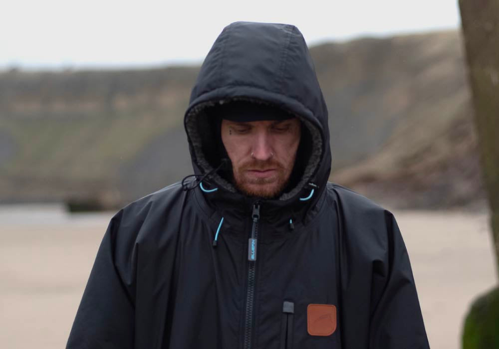 Person wearing a black hooded jacket, looking down on a beach with a blurred background.