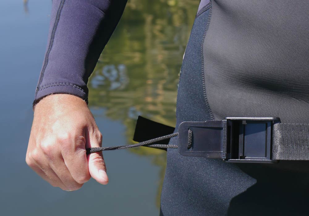 Person in a wetsuit pulling a strap on a paddleboard near calm water