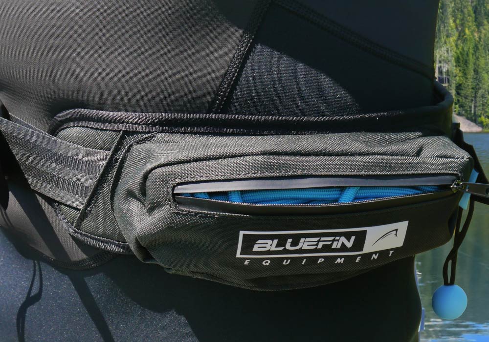 Black waist pouch with zipper, logo, and blue interior, worn on a wetsuit by water.