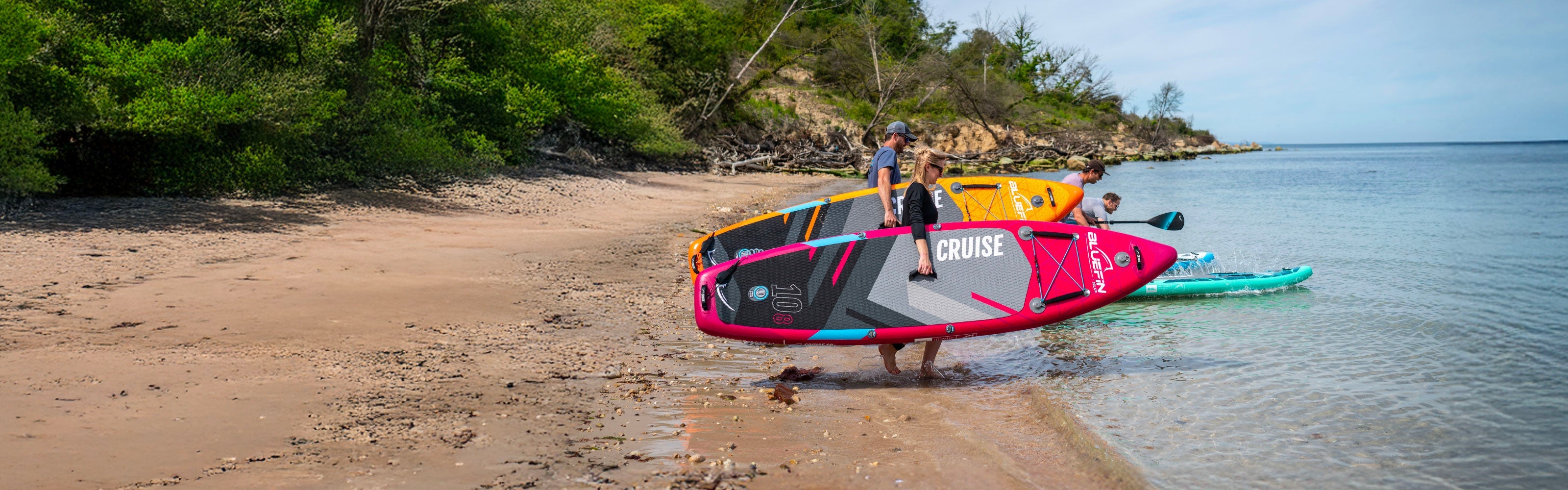 Two inflatable paddleboards in pink and orange on a sandy beach with people carrying them.