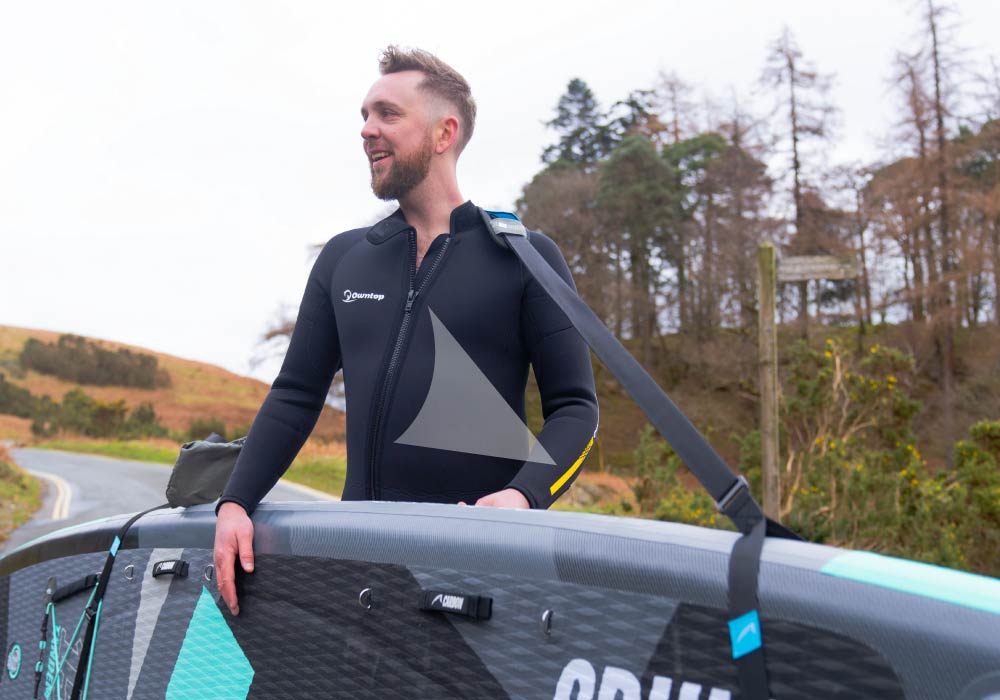 Man in a black wetsuit holding a paddleboard with a textured deck pad and fin, standing outdoors.