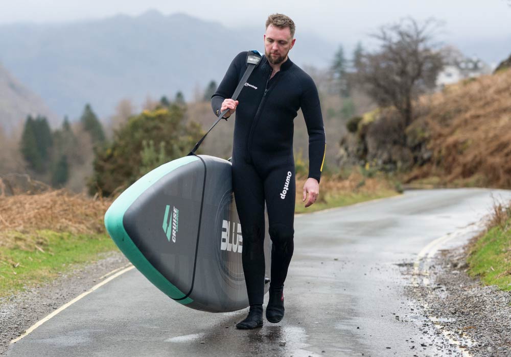 Man in a wetsuit carrying a paddleboard along a rural road with hills in the background.