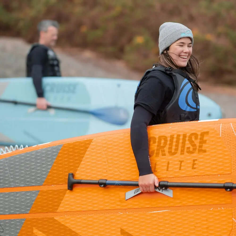 Deux personnes à la plage avec une planche de paddle orange et une planche de paddle bleu clair, souriant et se préparant à pagayer.