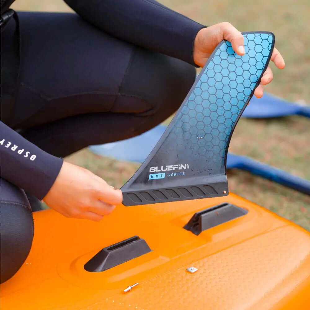 Personne tenant une dérive hexagonale bleue étiquetée BLUEFIN NXT SERIES au-dessus d'un paddleboard orange.