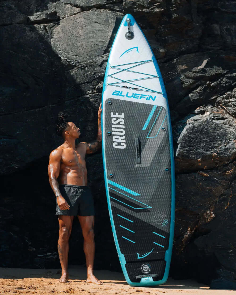 Man standing next to Bluefin Cruise inflatable paddleboard on sandy beach with rocky background