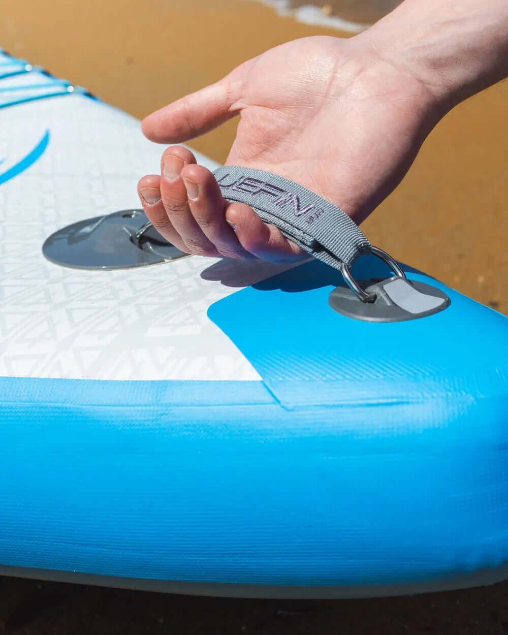 Hand holding the gray strap of a blue inflatable paddleboard on sandy beach
