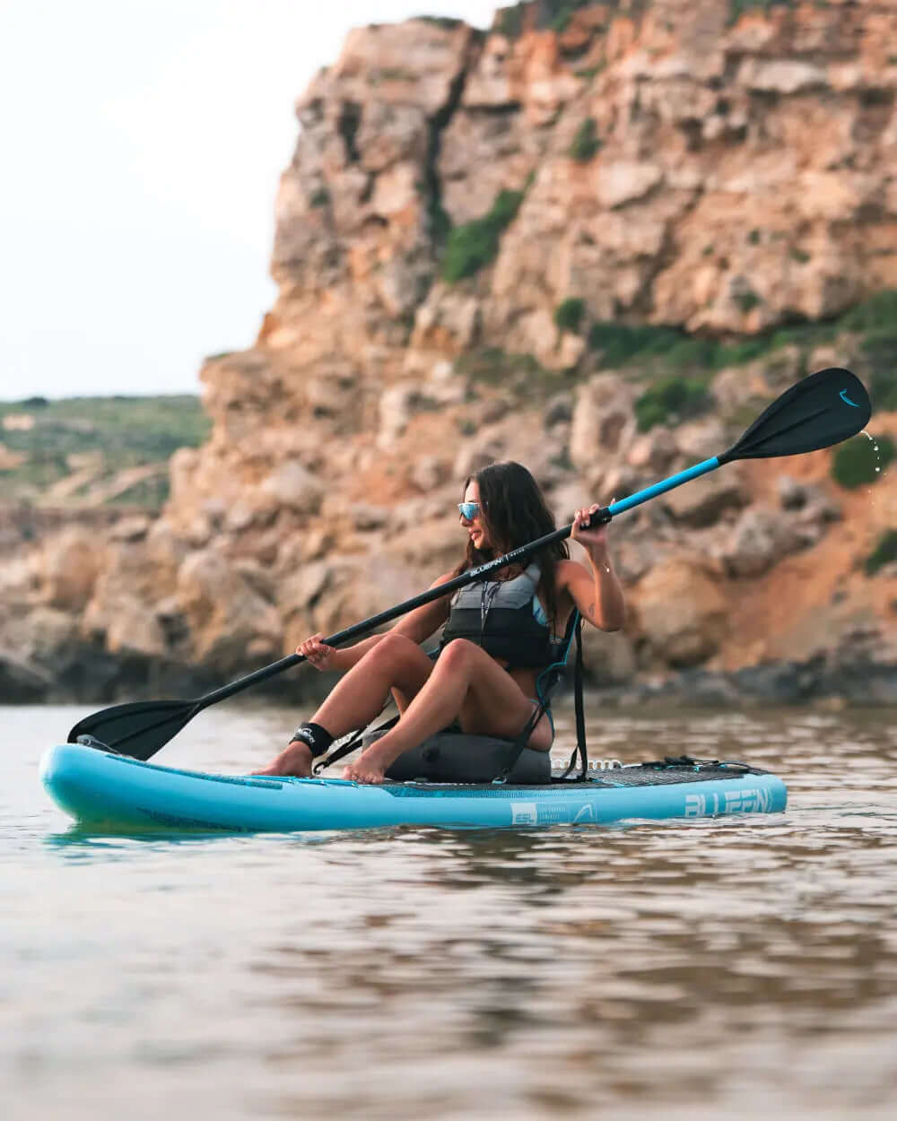 Woman paddling on a blue inflatable paddleboard near rocky cliffs in calm water
