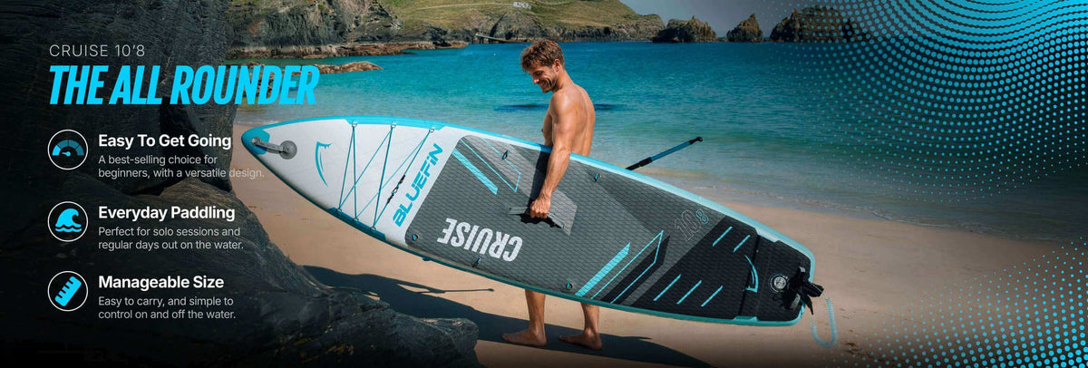 Mann hält ein aufblasbares Bluefin Cruise 10'8 Paddleboard an einem Strand mit felsigem Hintergrund