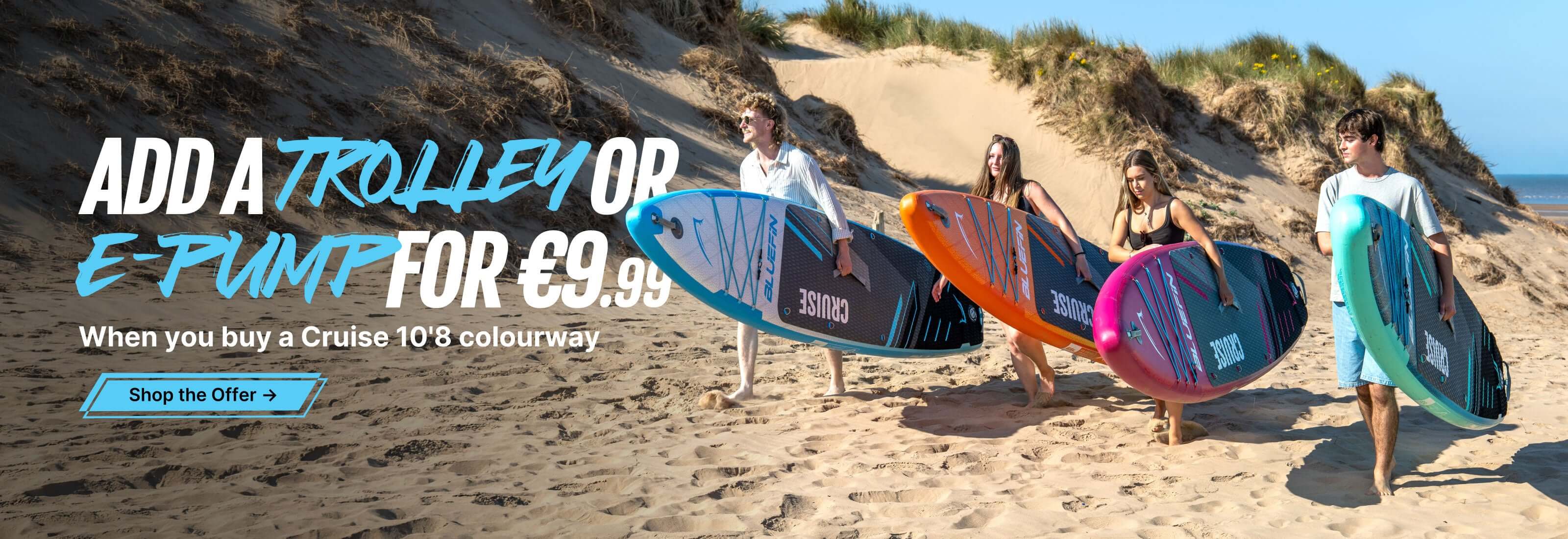 Group of four individuals carrying colorful stand-up paddleboards on a sandy beach under clear blue skies.