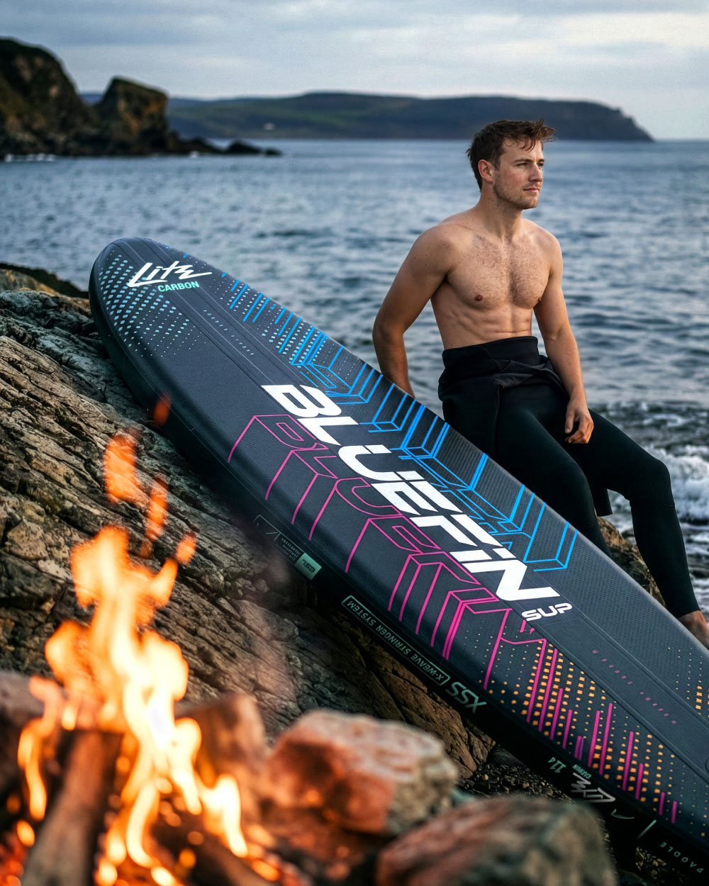 Man sitting on rocks by the water with a Bluefin SUP paddleboard and a campfire nearby.