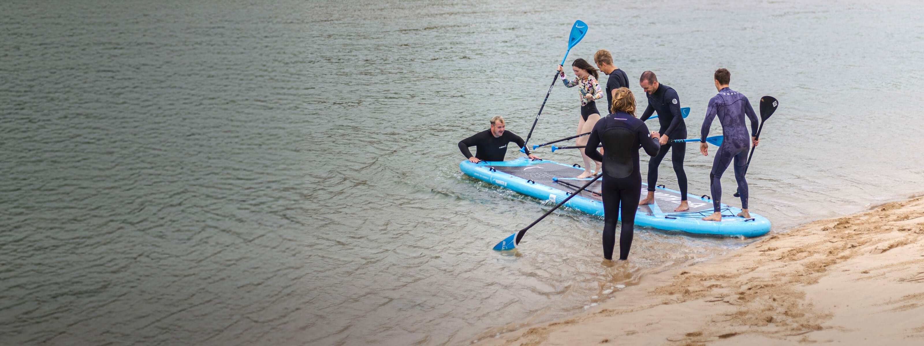 Group of six people on a large blue inflatable paddleboard in shallow water, some paddling and others balancing.