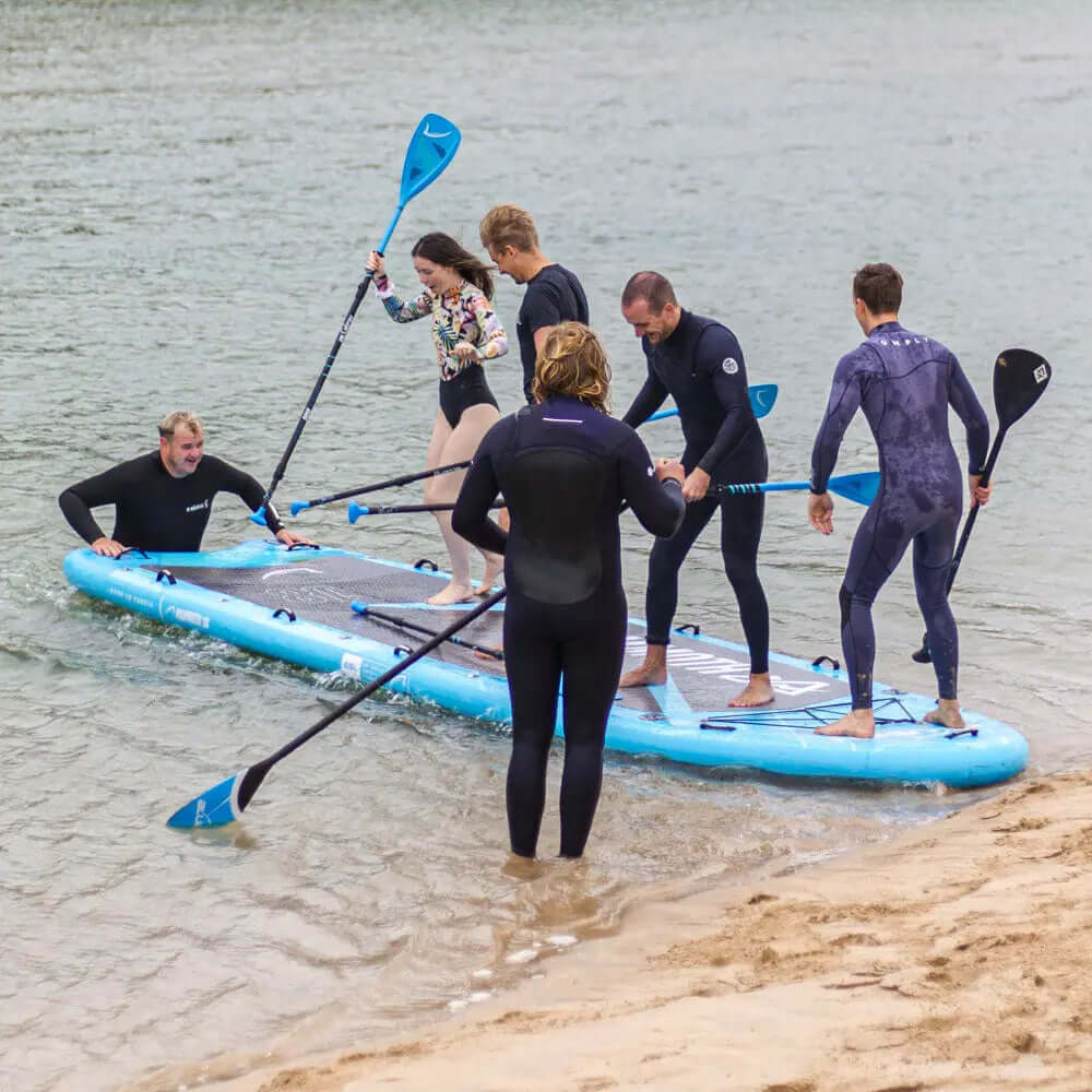 Gruppe von sechs Personen auf einem großen blauen aufblasbaren Paddleboard im flachen Wasser, einige paddeln, andere balancieren.