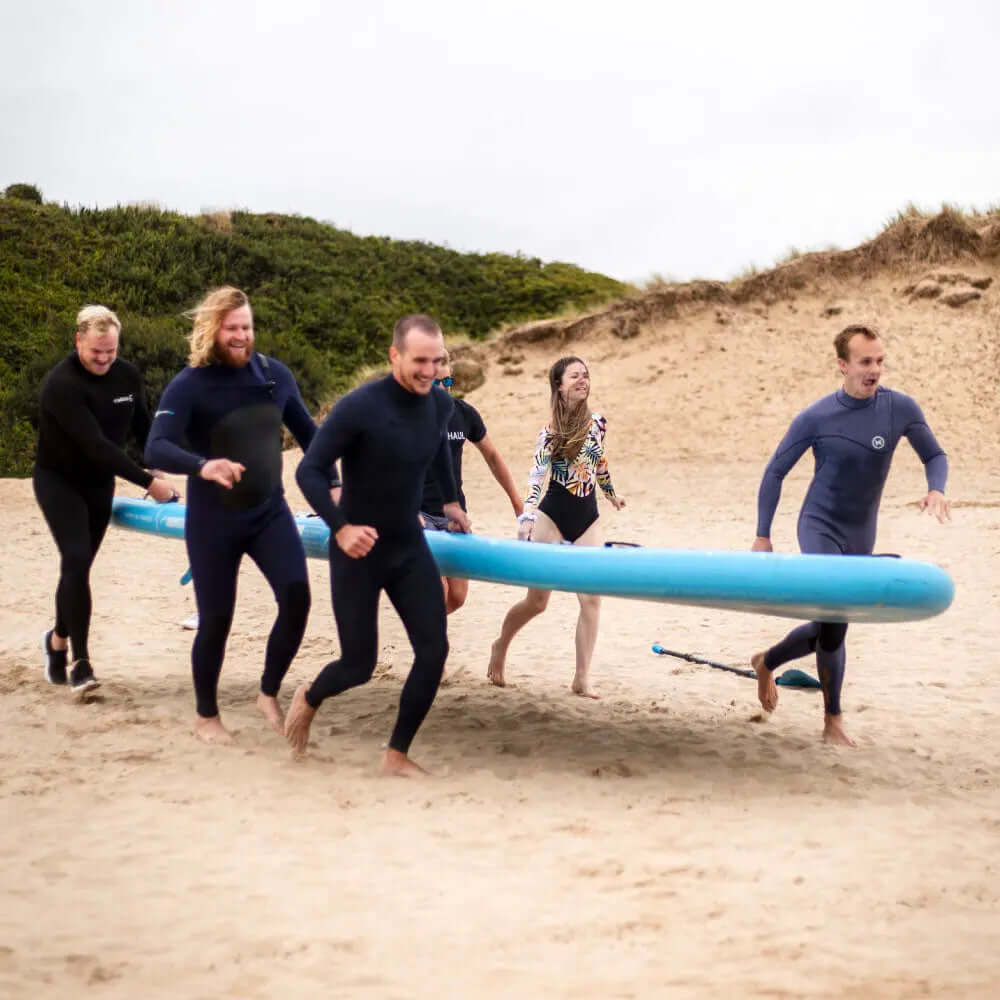 Gruppe von sechs Personen in Neoprenanzügen, die mit einem großen blauen Paddleboard über den Sand laufen