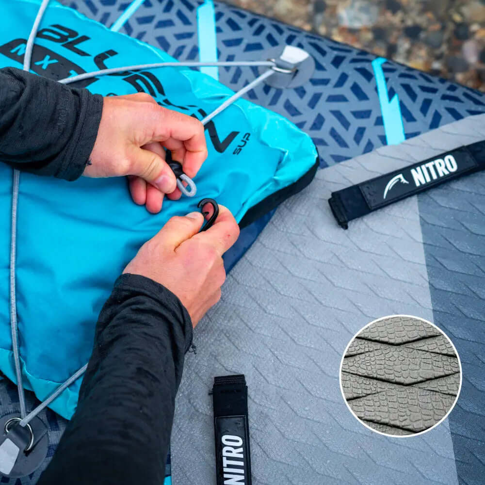Mãos segurando uma bolsa de prancha de paddle inflável azul com um deck pad texturizado cinza e strap NITRO.