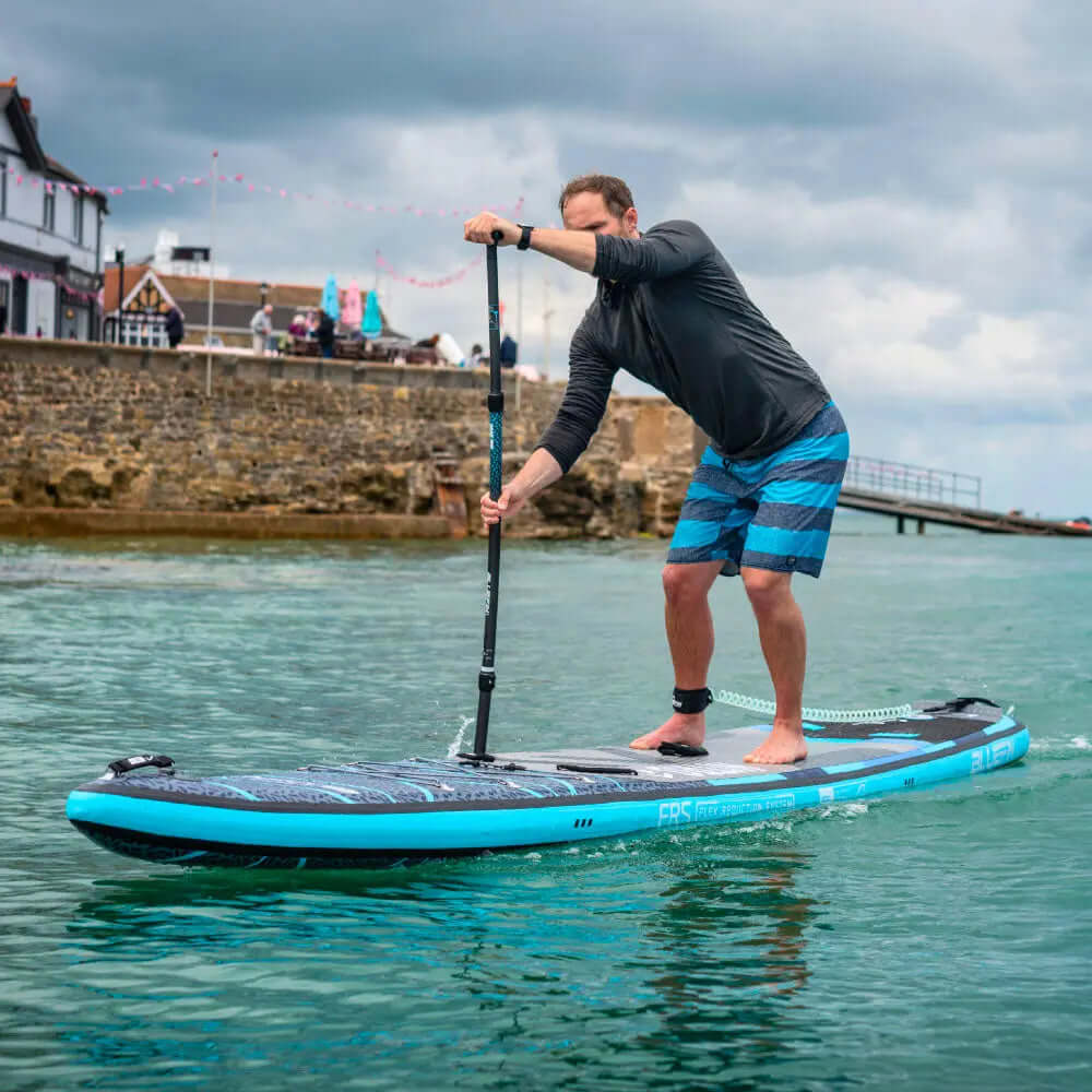 Hombre remando en una tabla de paddleboard inflable azul en agua tranquila, vistiendo camiseta negra y pantalones cortos a rayas.