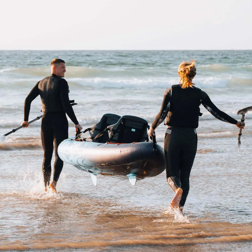 Two people in wetsuits carrying an inflatable paddleboard through shallow water at the beach