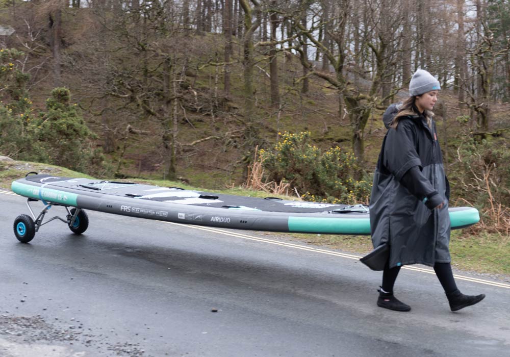 Person in a long coat walking with a wheeled inflatable paddleboard on a road