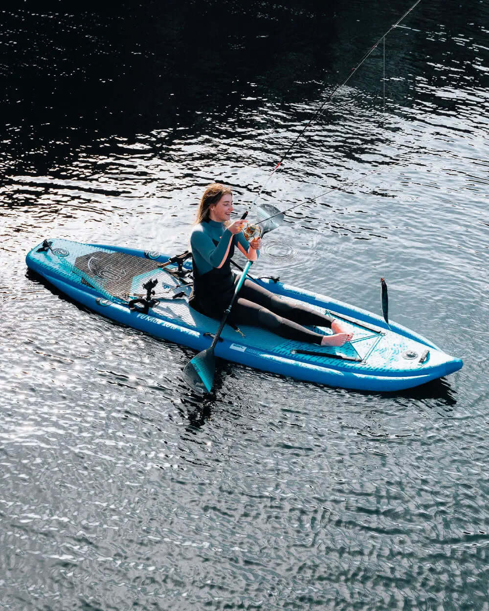 Woman fishing from a blue inflatable paddleboard on calm water, wearing a wetsuit.