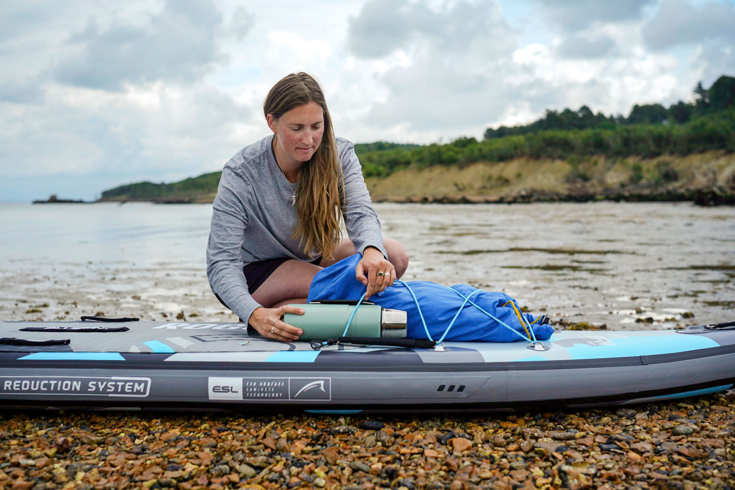 Woman securing a blue bag and thermos on an inflatable paddleboard by the shore