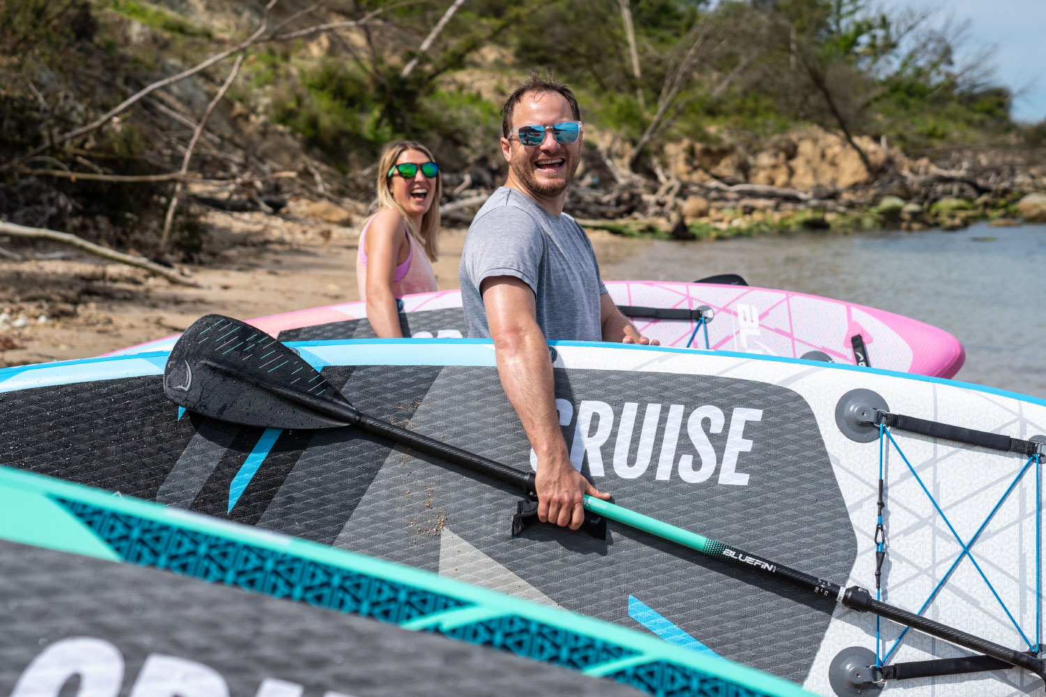 Two people smiling near inflatable paddleboards on a beach, one holding a paddle.