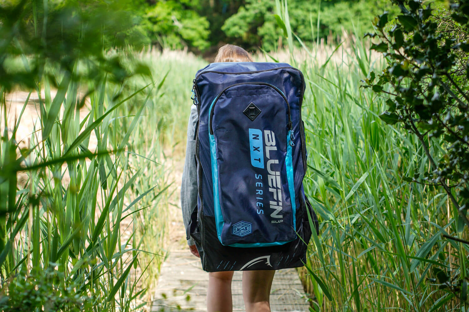 Person walking on a wooden path carrying a large blue backpack with Bluefin branding in green and white