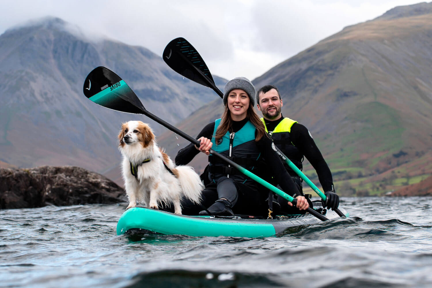 Two people and a dog on a teal inflatable paddleboard, paddling in a lake with mountains in the background.
