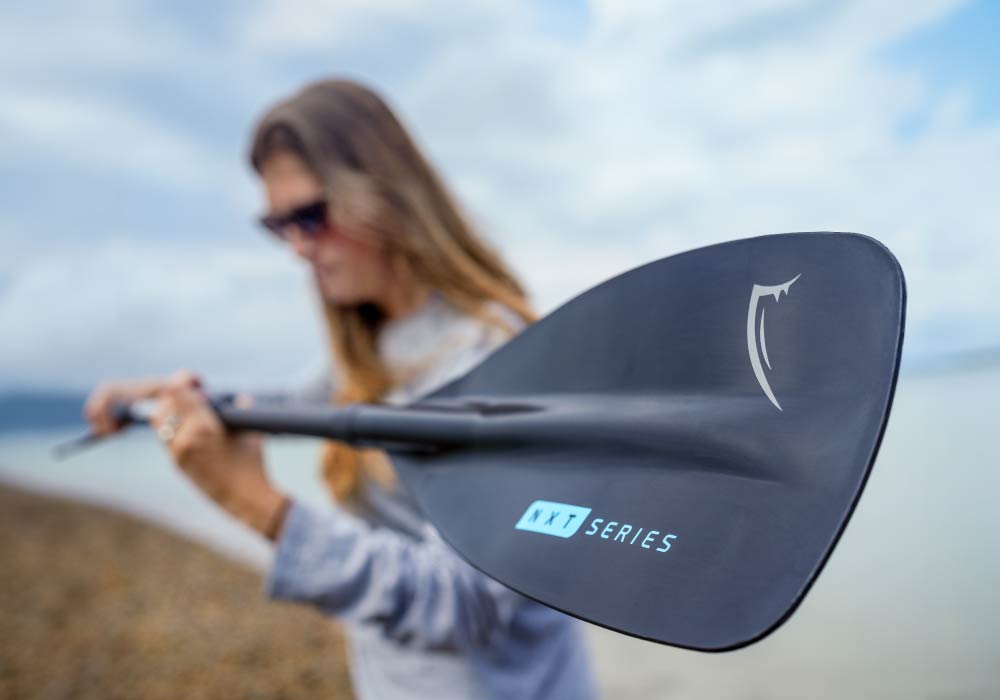 Gros plan sur une pale de pagaie noire avec le texte 'NXT SERIES', tenue par une personne sur une plage.