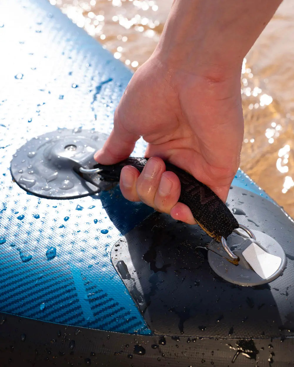 Hand gripping the handle of a blue inflatable paddleboard with water droplets on the surface