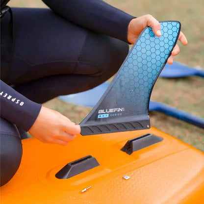 Person hält eine blaue hexagonale Finne mit der Aufschrift BLUEFIN NXT SERIES über einem orangefarbenen Paddleboard.