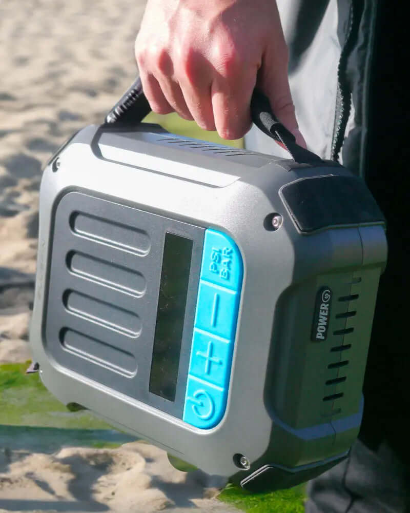 Tragbare Luftpumpe mit blauem Bedienfeld, wird an einem sandigen Strand gehalten.