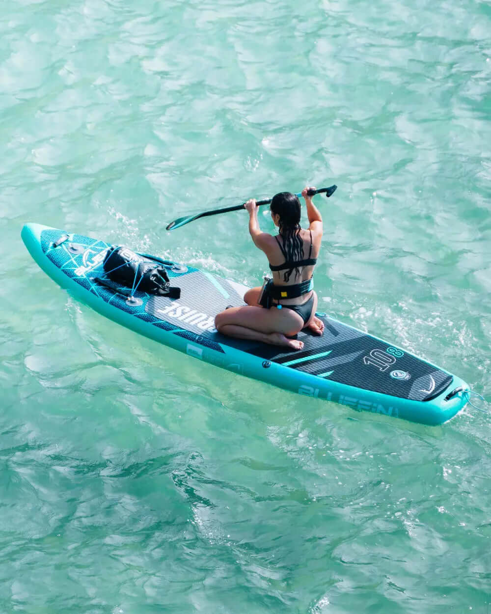 Person kneeling on a blue inflatable paddleboard, paddling in clear turquoise water.