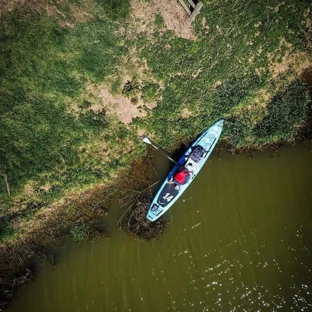 Vue aérienne d'un kayak bleu sur une berge verte avec une personne en train de pagayer et de l'herbe entourant l'eau.