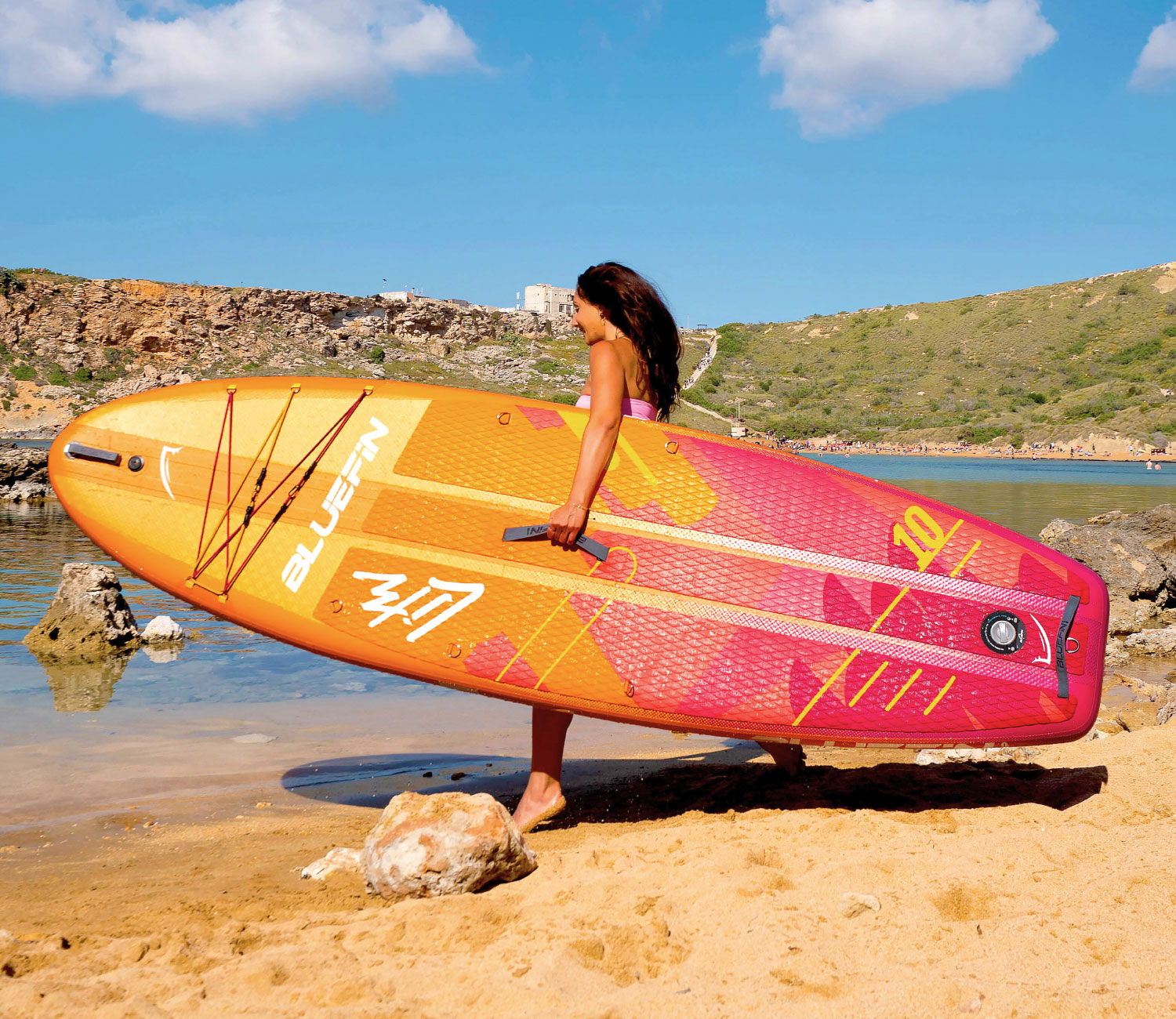 Person carrying a colorful inflatable paddleboard on a sandy beach near water.