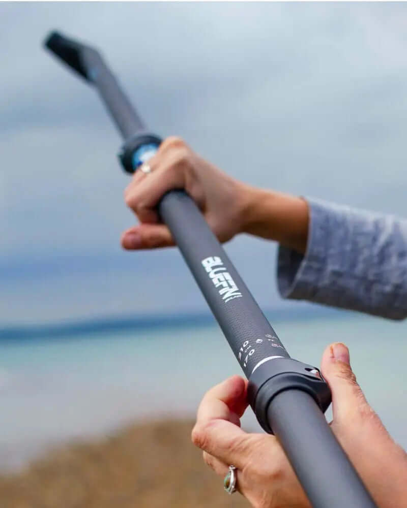 Personne tenant une pagaie ajustable noire avec une prise texturée et une extrémité courbée, contre un ciel nuageux.