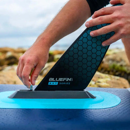 Person, die eine schwarze sechseckige Finne an einem Bluefin Paddleboard an einer felsigen Küste anbringt.