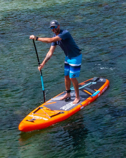 Man paddling on an orange inflatable paddleboard in clear water, wearing blue shorts and sunglasses.