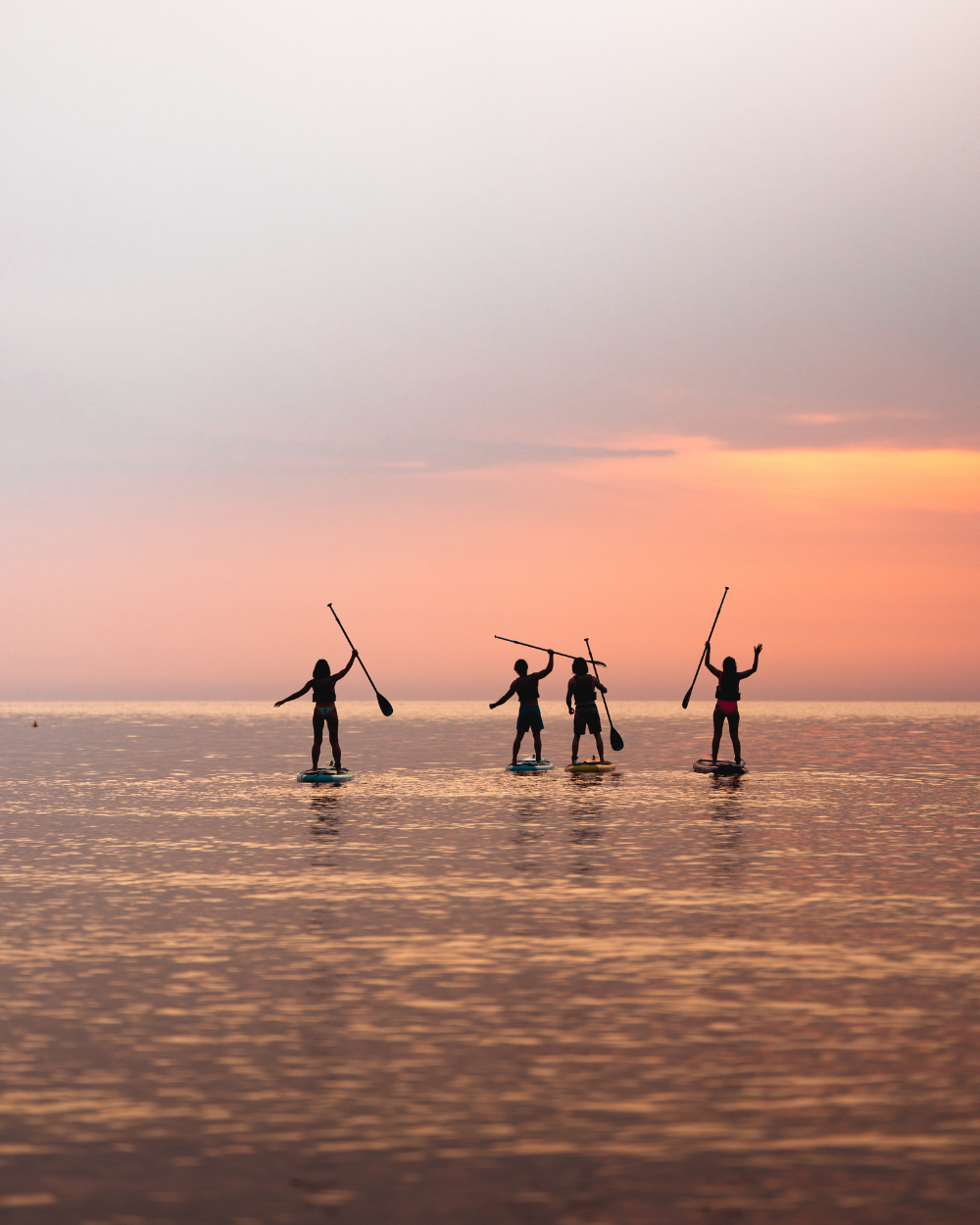 Four people paddleboarding at sunset, silhouetted against a colorful sky on calm water.