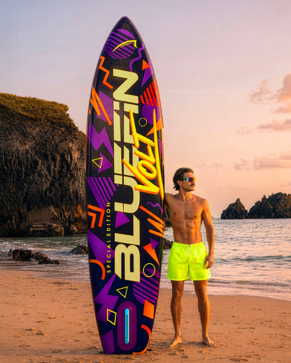Colorful inflatable paddleboard with geometric designs, man in neon shorts standing on beach at sunset